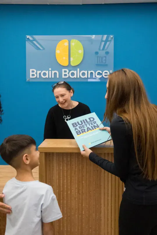 A front desk hand-off of a brochure to a parent, with a boy standing by