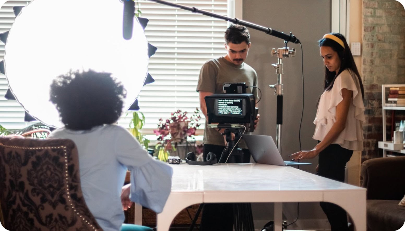 Sociallyin Studios crew prepares a video shoot with seated talent, a camera and teleprompter, a large studio light, and a boom microphone.